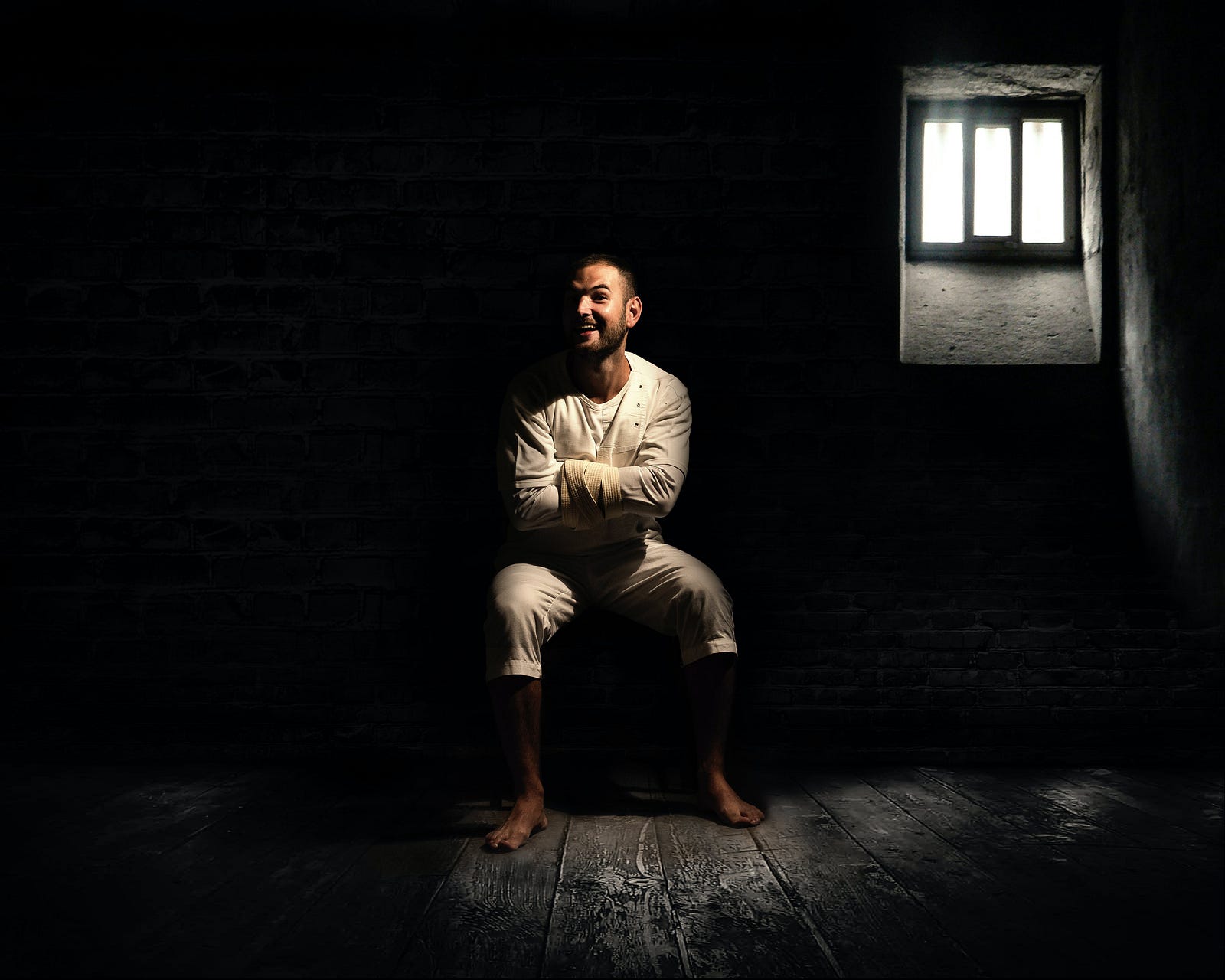 man in straight jacket and laughs to himself with a disturbed look on his face in a darkened room with one window and a small amount of light shining through the window