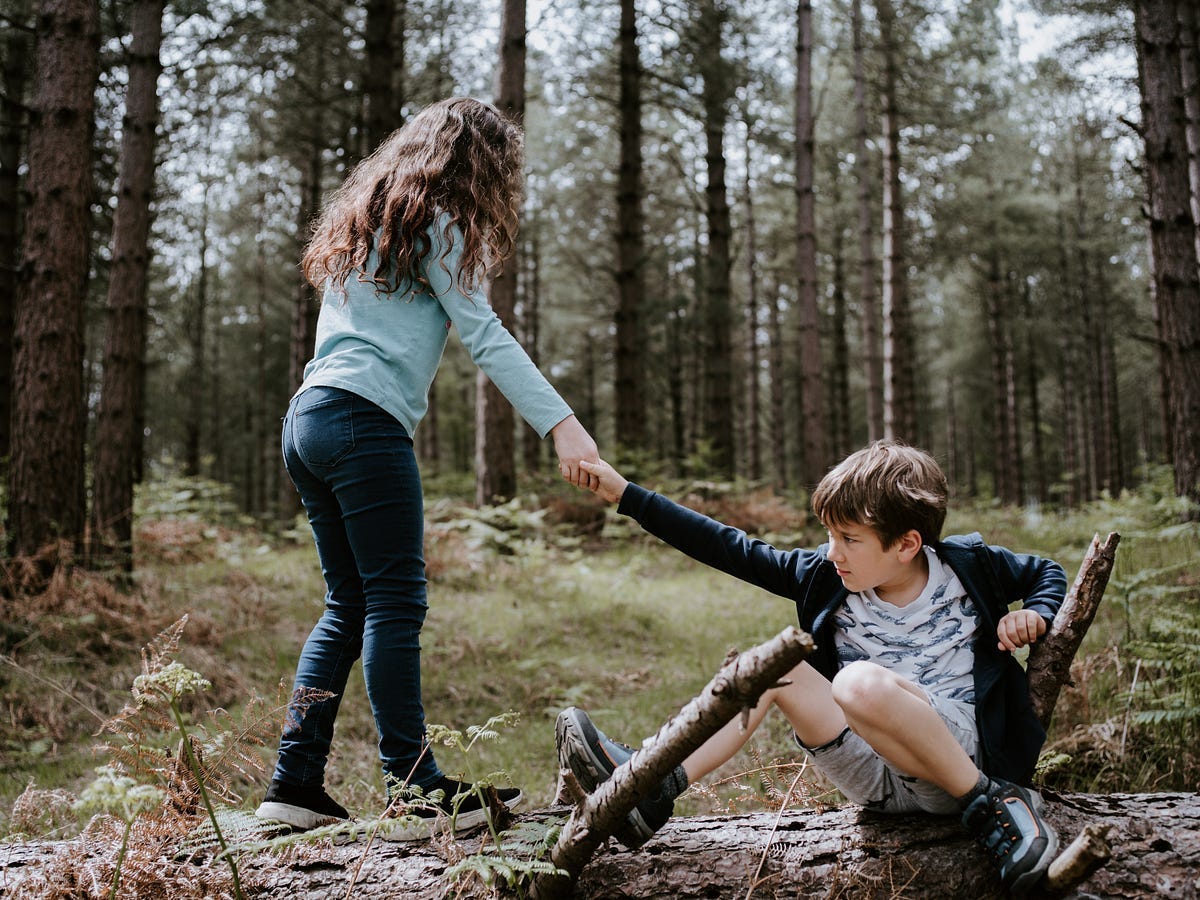 A child is helping another child stand up. They are in a forest.