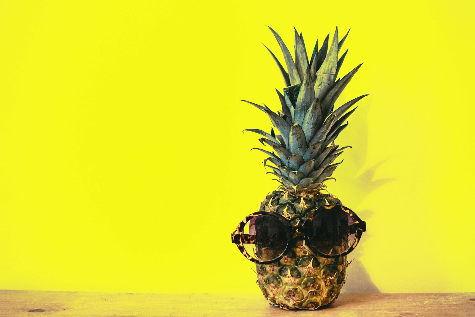 a pineapple wearing sunglasses, placed on a wooden table in front of a yellow wall